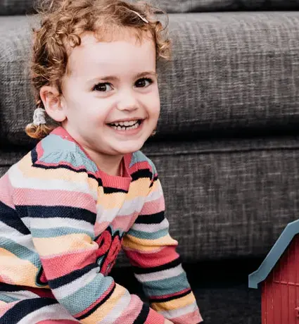 A little girl is smiling while playing with toys and sitting on the floor near a grey sofa.