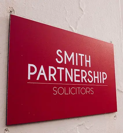 The front of the Stoke Smith Partnership office building, focussed on a red sign saying 'Smith Partnership'. The front of the building is cream, and there is a tree visible down the road nearby.